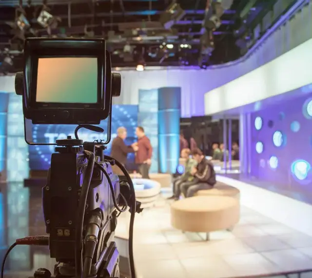 Telecamera in primo piano in uno studio televisivo. Si intravede il backstage, con persone sedute e in piedi, luci soffuse e un'atmosfera di attesa.
