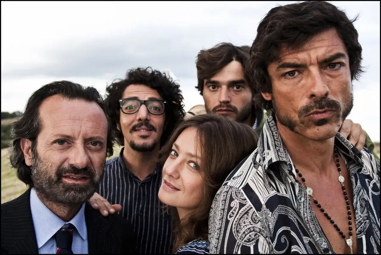 Un gruppo di attori posa per un film di commedie comiche. Un uomo con la barba guarda dritto, mentre una donna sorride.