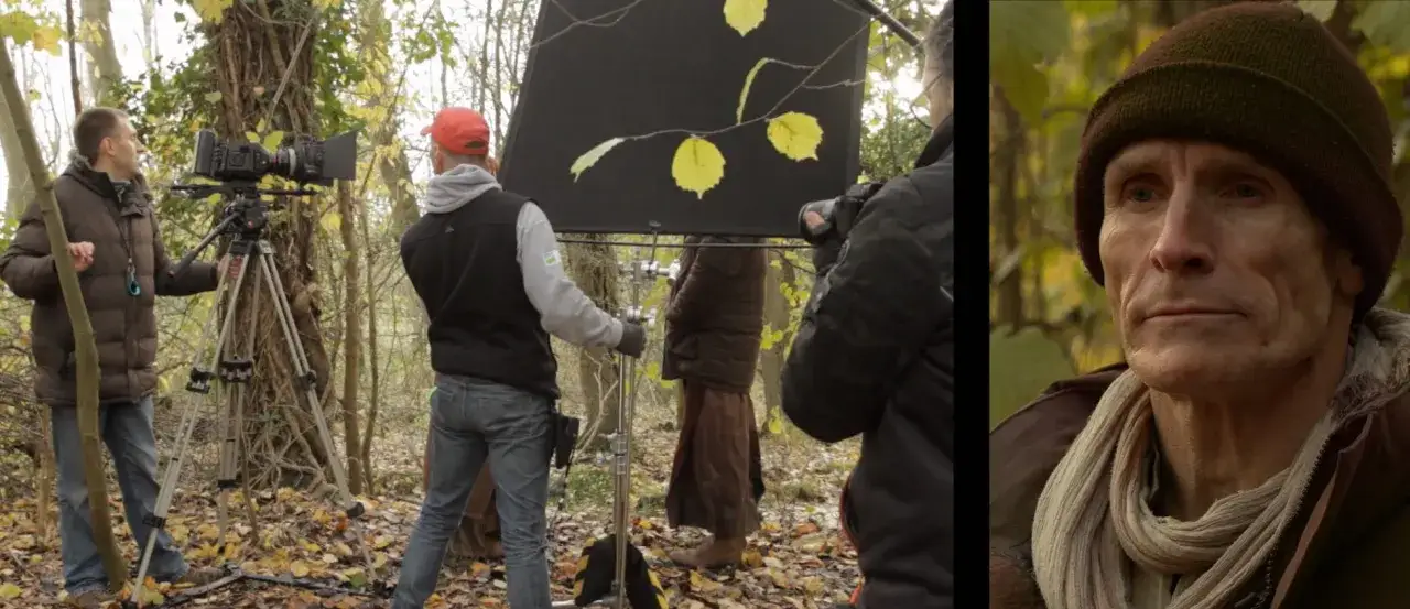 Una foto di scena mostra un team di ripresa in un bosco autunnale. Un operatore dirige la telecamera, mentre un altro assistente regola un pannello riflettente. In primo piano, un uomo con un berretto e una sciarpa guarda pensieroso.