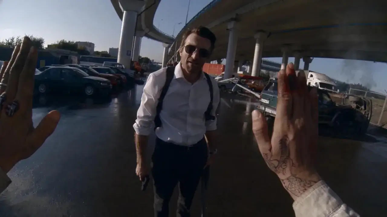 Un uomo con occhiali da sole e camicia bianca, armato, si trova in un parcheggio disordinato. &Egrave; il narratore protagonista di questa scena caotica.