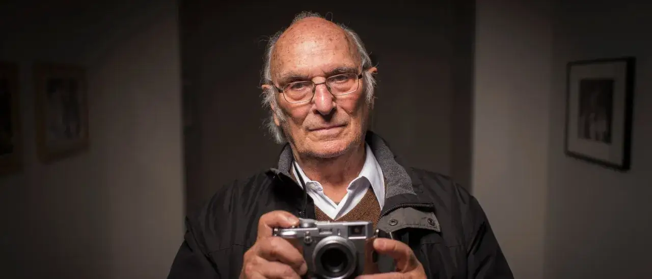 Un anziano regista spagnolo con occhiali tiene una fotocamera, con quadri alle pareti.
