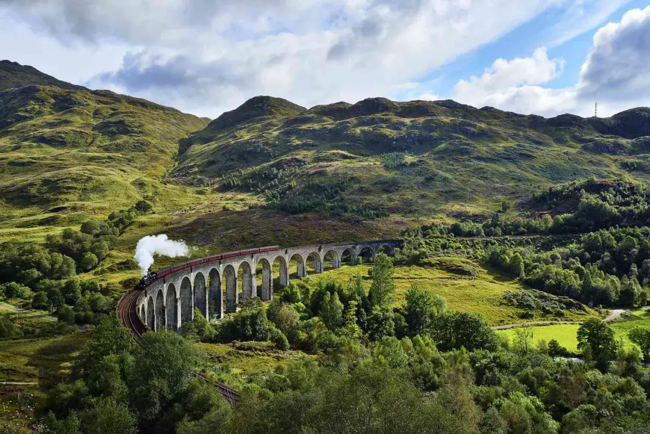 Treno a vapore attraversa un viadotto in Scozia, evocando scene di film epici tra montagne verdi e cielo nuvoloso.
