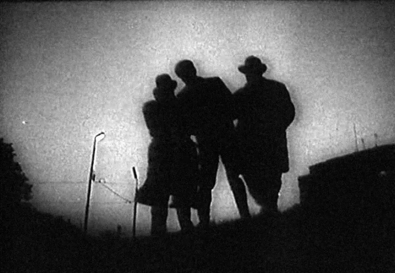 Tre figure in silhouette, forse parte de il processo Welles, si stagliano contro un cielo nebbioso, con un lampione a sinistra.