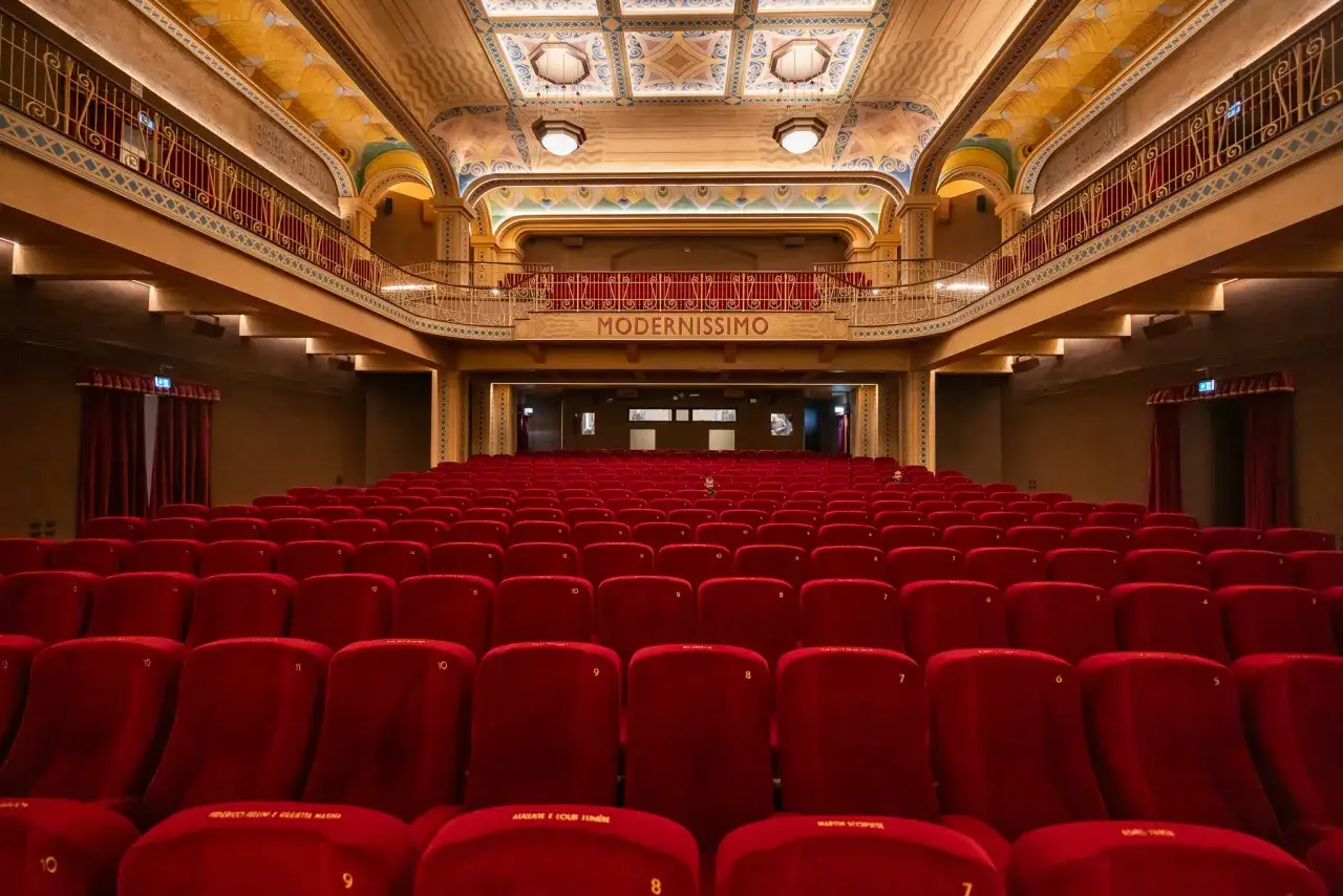 Interno di un cinema con file di poltrone rosse vellutate. Sopra, la balconata e il soffitto decorato. Si intravede la scritta 
