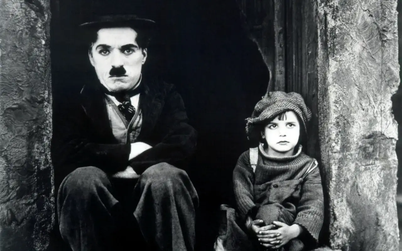 Charlie Chaplin e un bambino, icone del film comico, siedono vicini in un'ambientazione scura.
