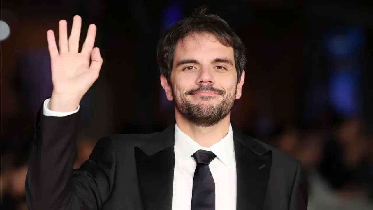 Valerio Vestoso saluta con la mano, sorridendo, vestito con un elegante abito scuro e cravatta.