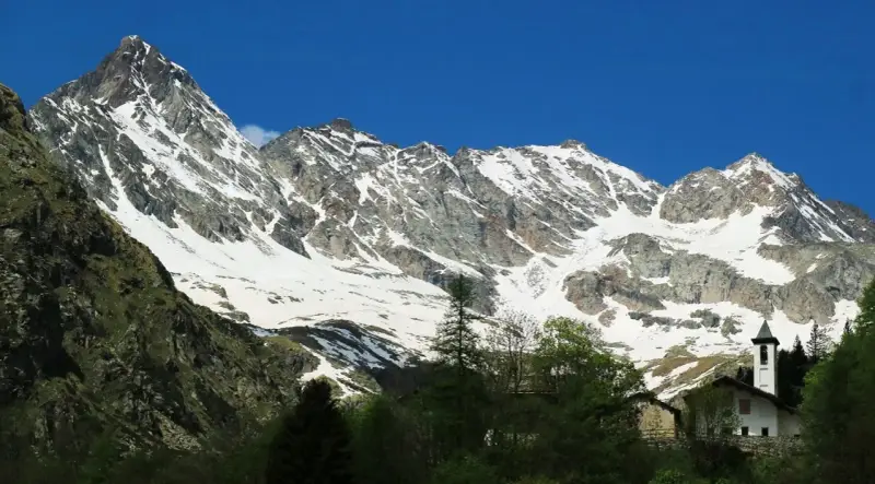 Imponente montagna sacra innevata, con un piccolo campanile bianco che emerge tra gli alberi verdi in primo piano.