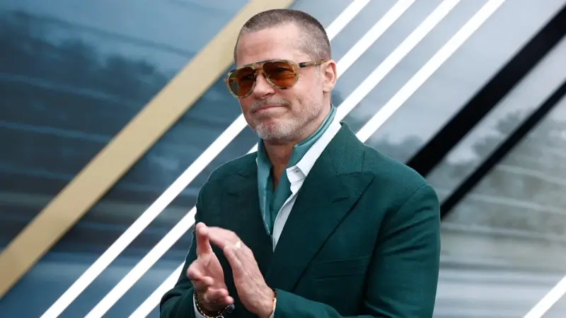 Brad Pitt, icona del cinema hollywoodiano, con occhiali da sole e completo verde, applaude con un sorriso.