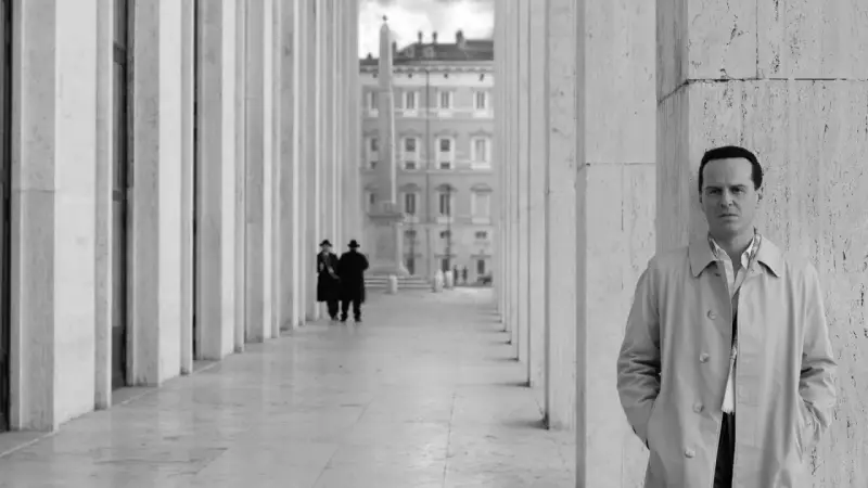 Un uomo in trench osserva l'obiettivo, mentre sullo sfondo due figure in abito scuro passeggiano sotto un colonnato. Potrebbe essere una scena da "Il talento di Mr. Ripley dove è stato girato".