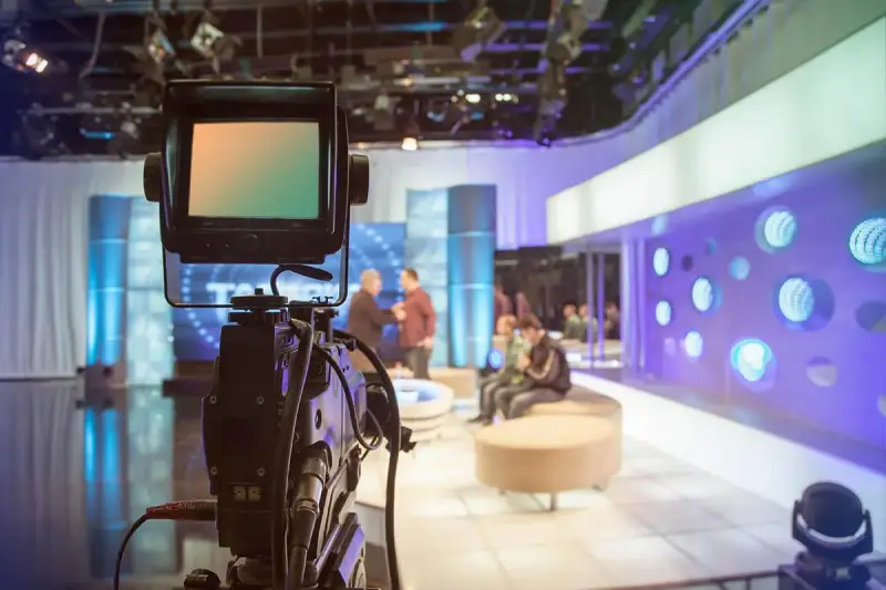 Telecamera in primo piano in uno studio televisivo. Si intravede il backstage, con persone sedute e in piedi, luci soffuse e un'atmosfera di attesa.